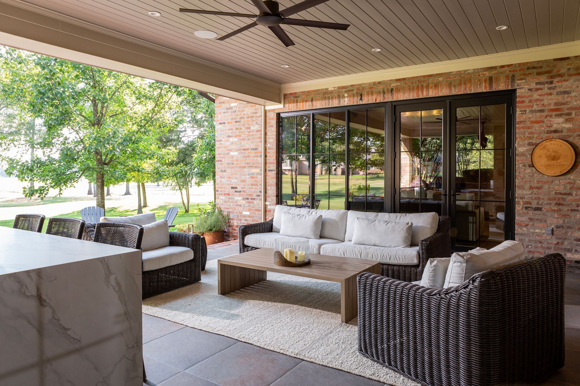 Covered patio with wicker seating, white sofas, coffee table, and brick wall overlooking a sunny yard