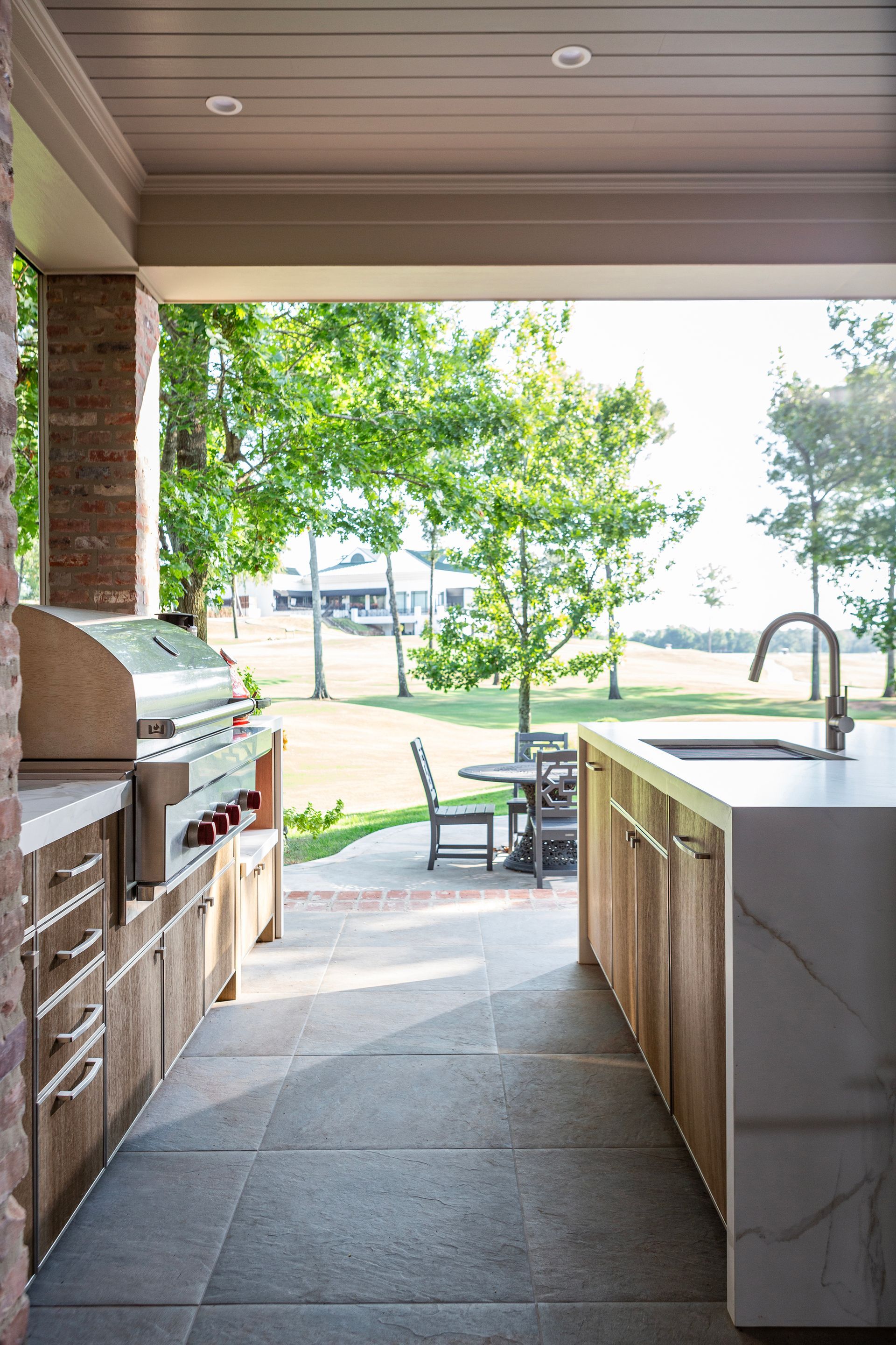 Covered patio with grill, sink island, and view of a sunny backyard with trees and patio furniture