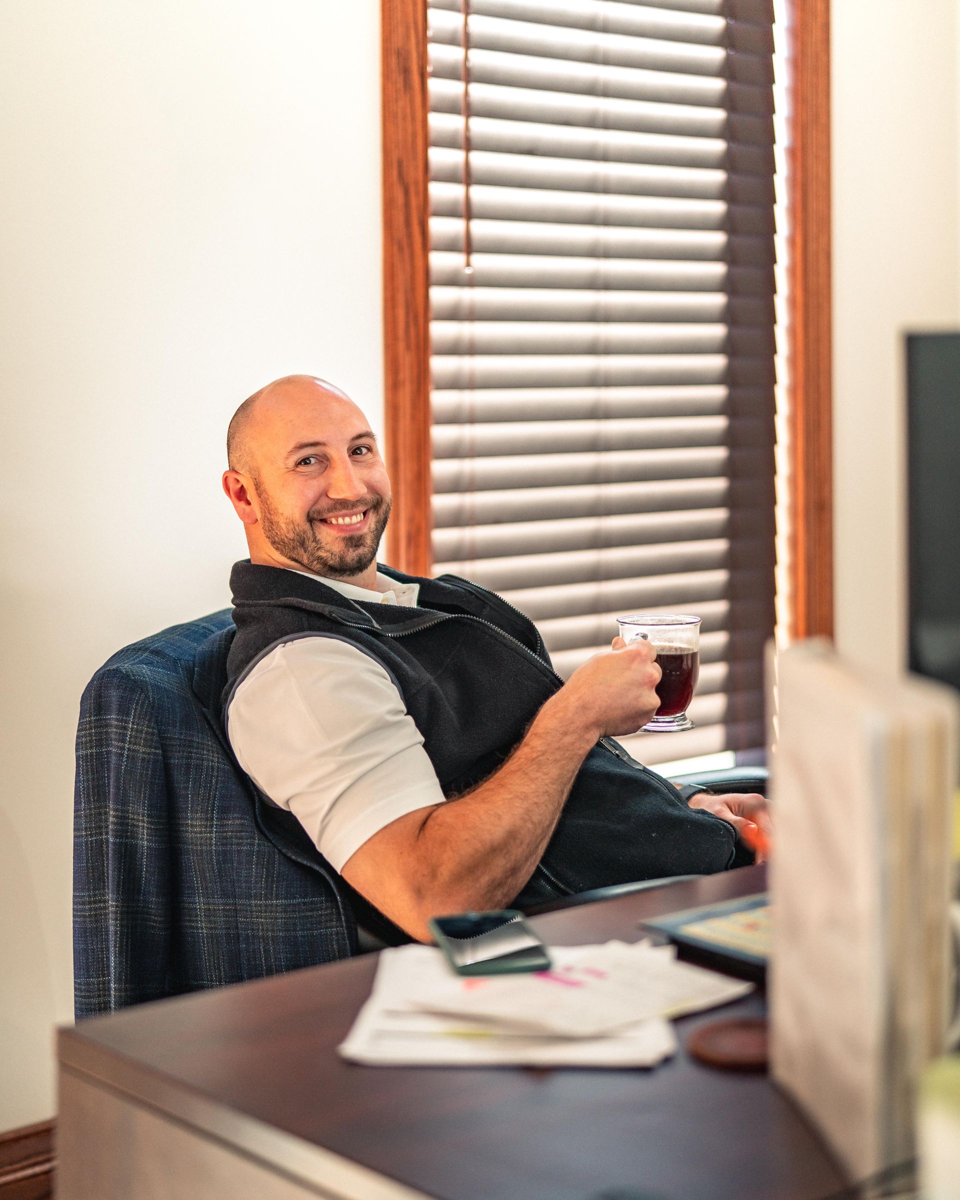 Smiling man at an office desk, holding a mug and looking toward the camera.