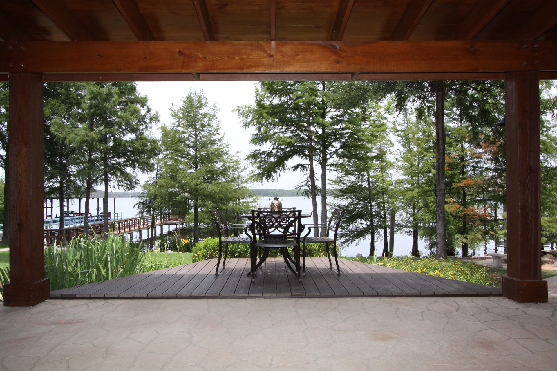 Covered patio overlooking a lake, with wooden chairs and a table framed by trees and pillars