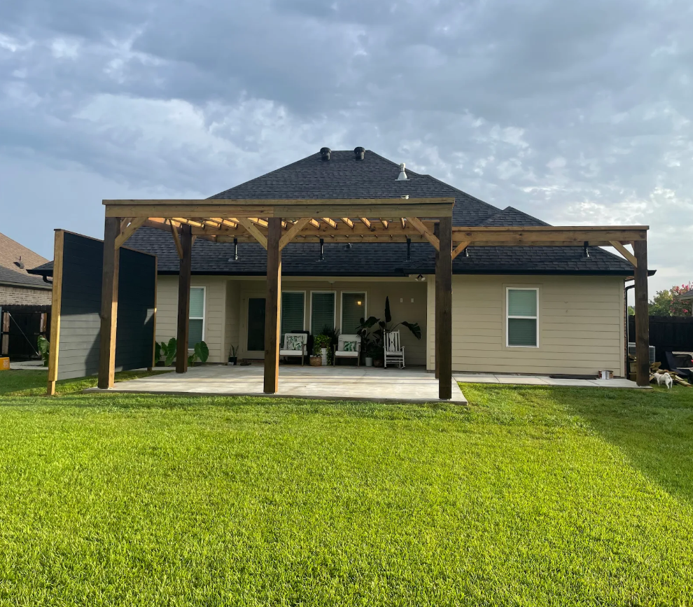 Backyard patio with wooden pergola, green lawn, and beige house under cloudy sky