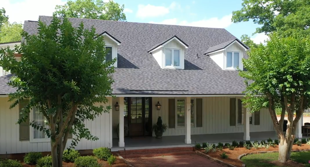 White house with gray roof and dormer windows, framed by trees and a front porch garden.