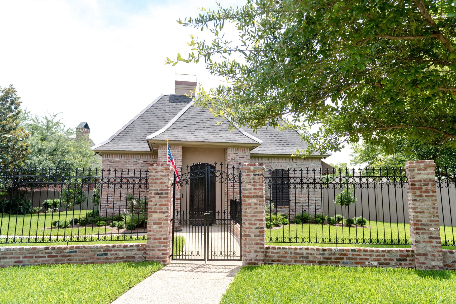 Brick house with black iron gate and fence, front yard, and large trees on a sunny day