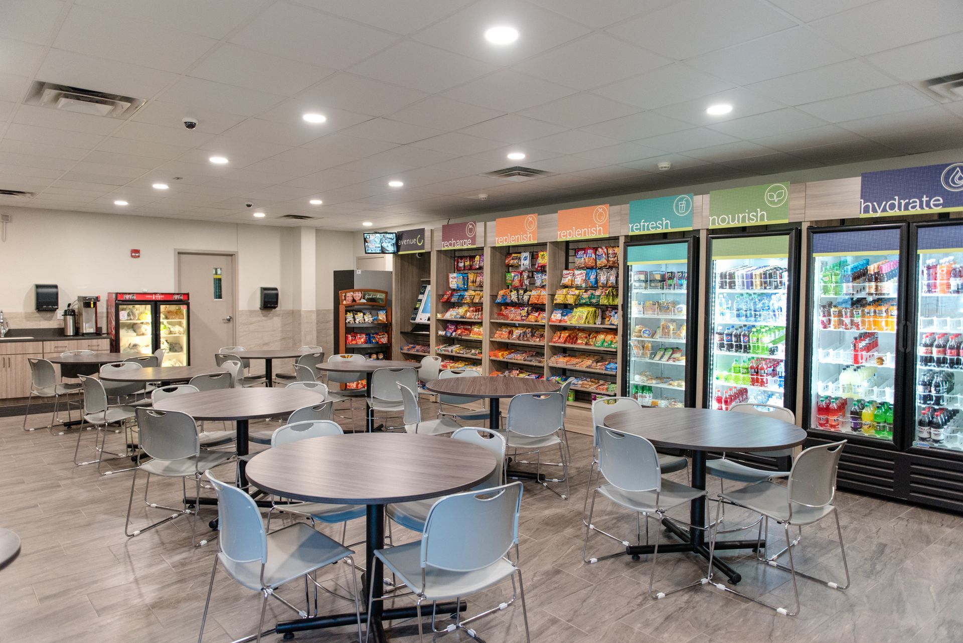 Empty cafeteria with round tables, gray chairs, and a snack and drink refrigerator wall.