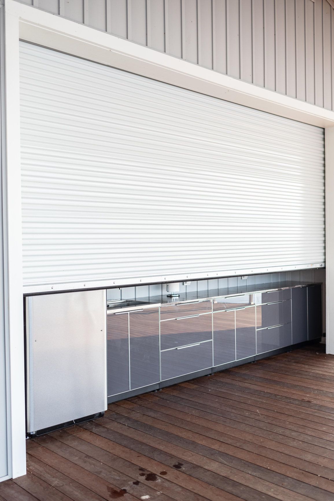 White roll-up garage door partially open over a wooden deck, showing a row of glass cabinets inside.