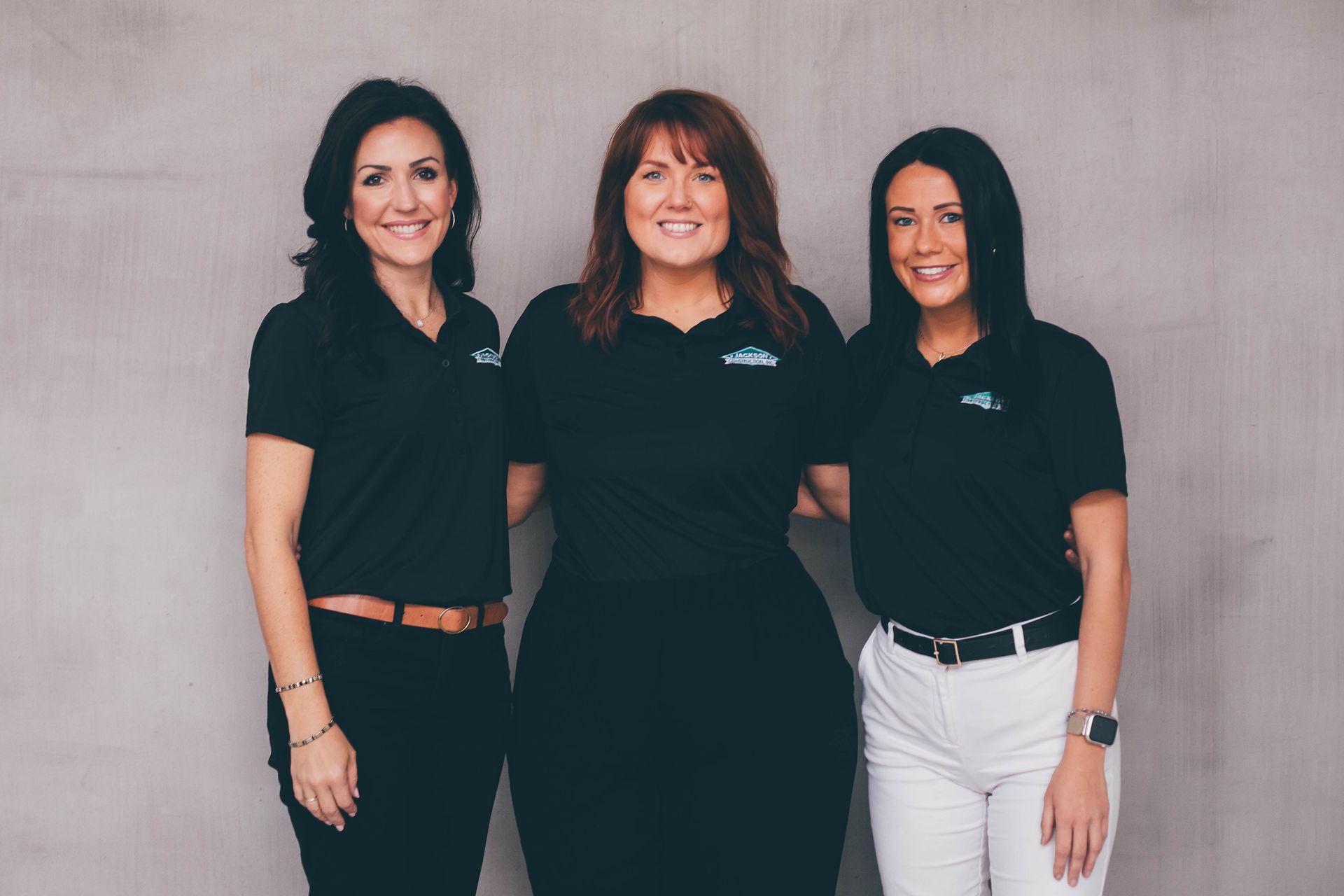 Three women in black polo shirts standing against a plain wall, smiling at the camera.