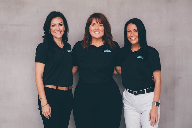 Three women in black polo shirts standing against a plain wall, smiling at the camera.