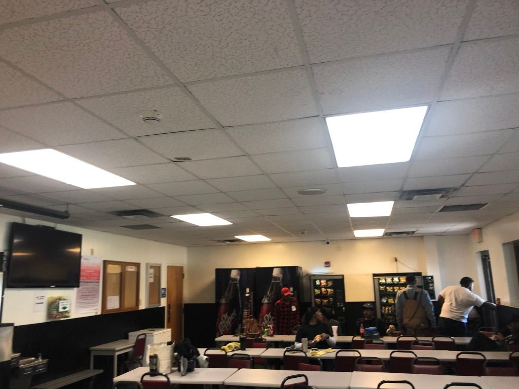 Classroom or meeting room with rows of chairs, desks, wall-mounted screens, and people at the back.