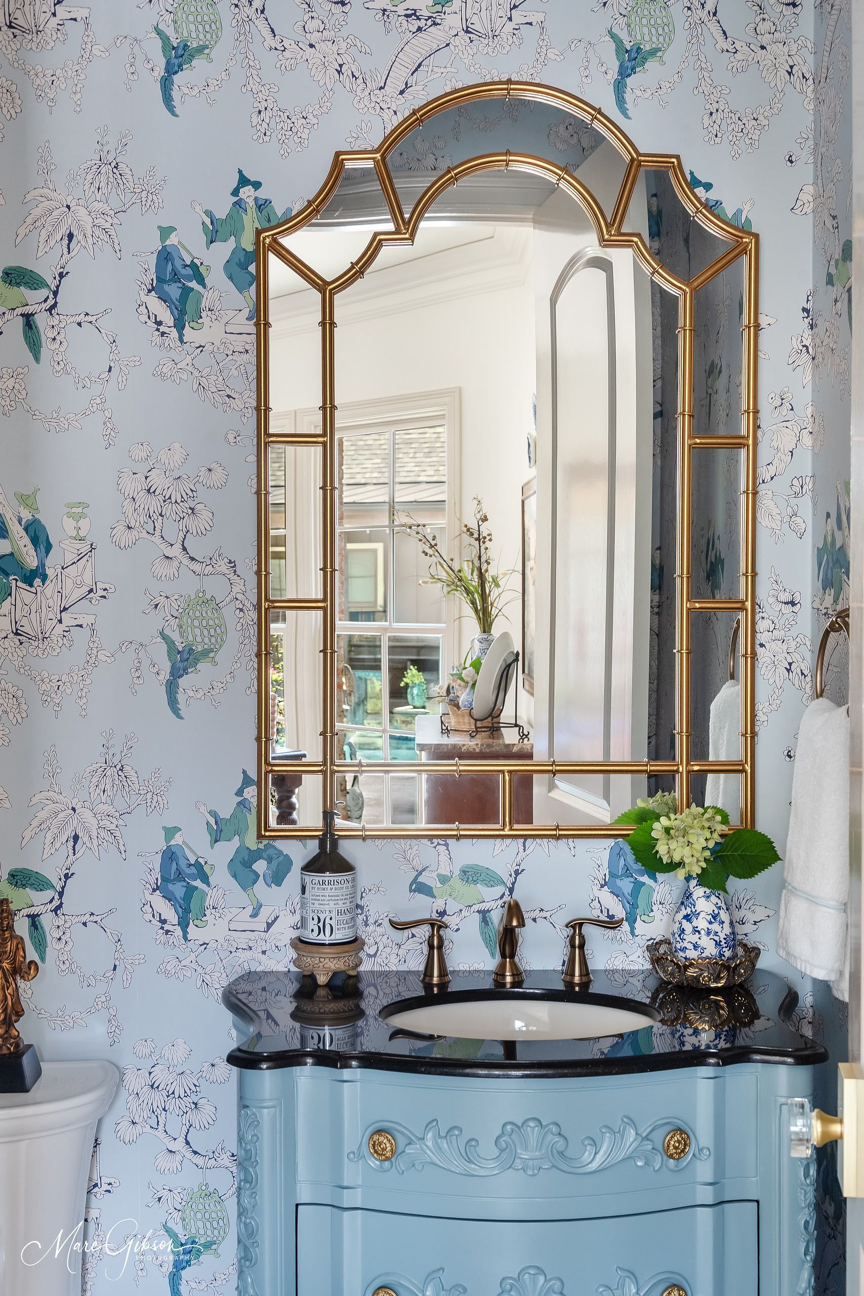 Blue floral bathroom vanity with gold-framed mirror and black countertop under a window.