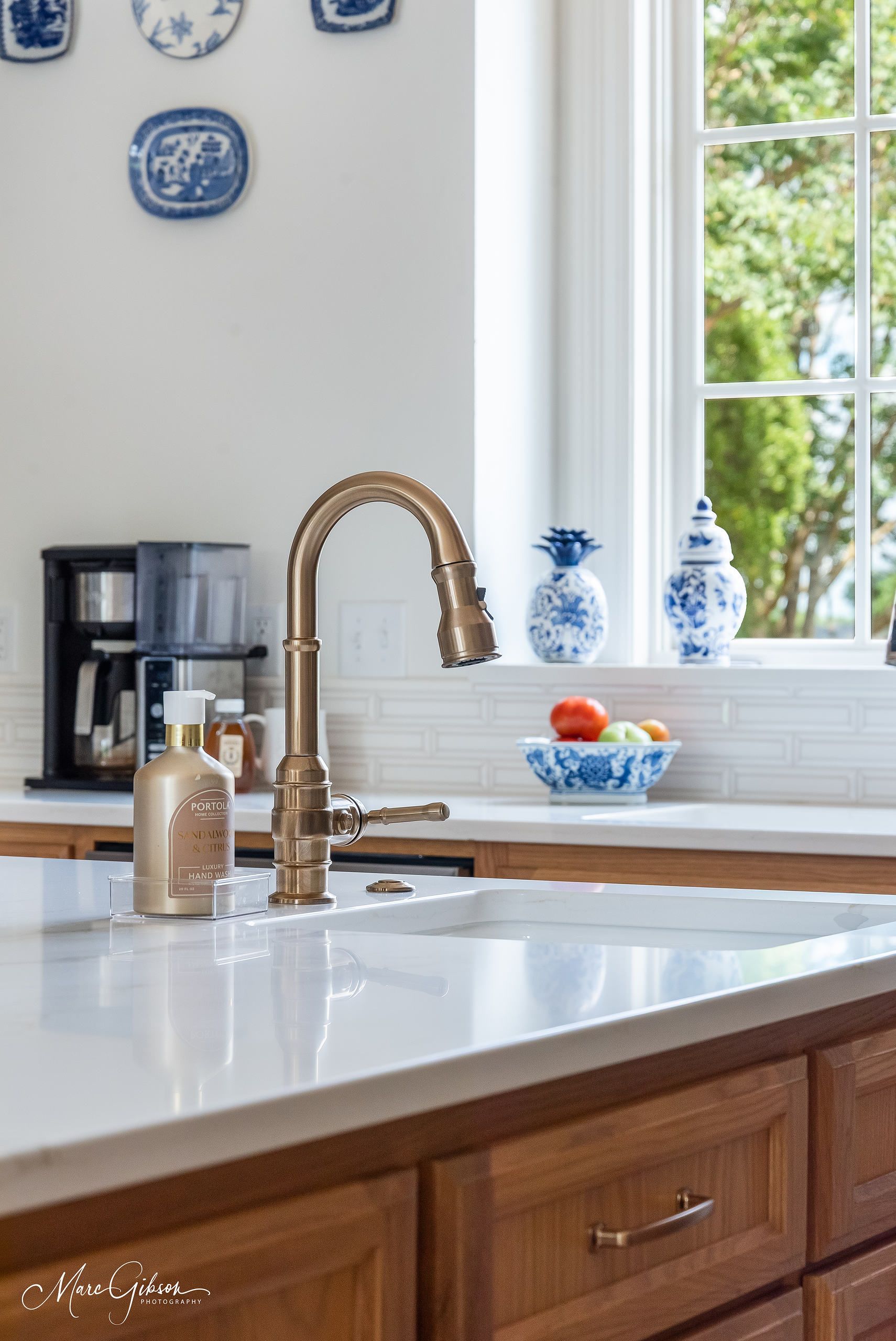 Bright kitchen sink with brass faucet, white countertop, wood cabinets, and window with blue decor and fruit bowl