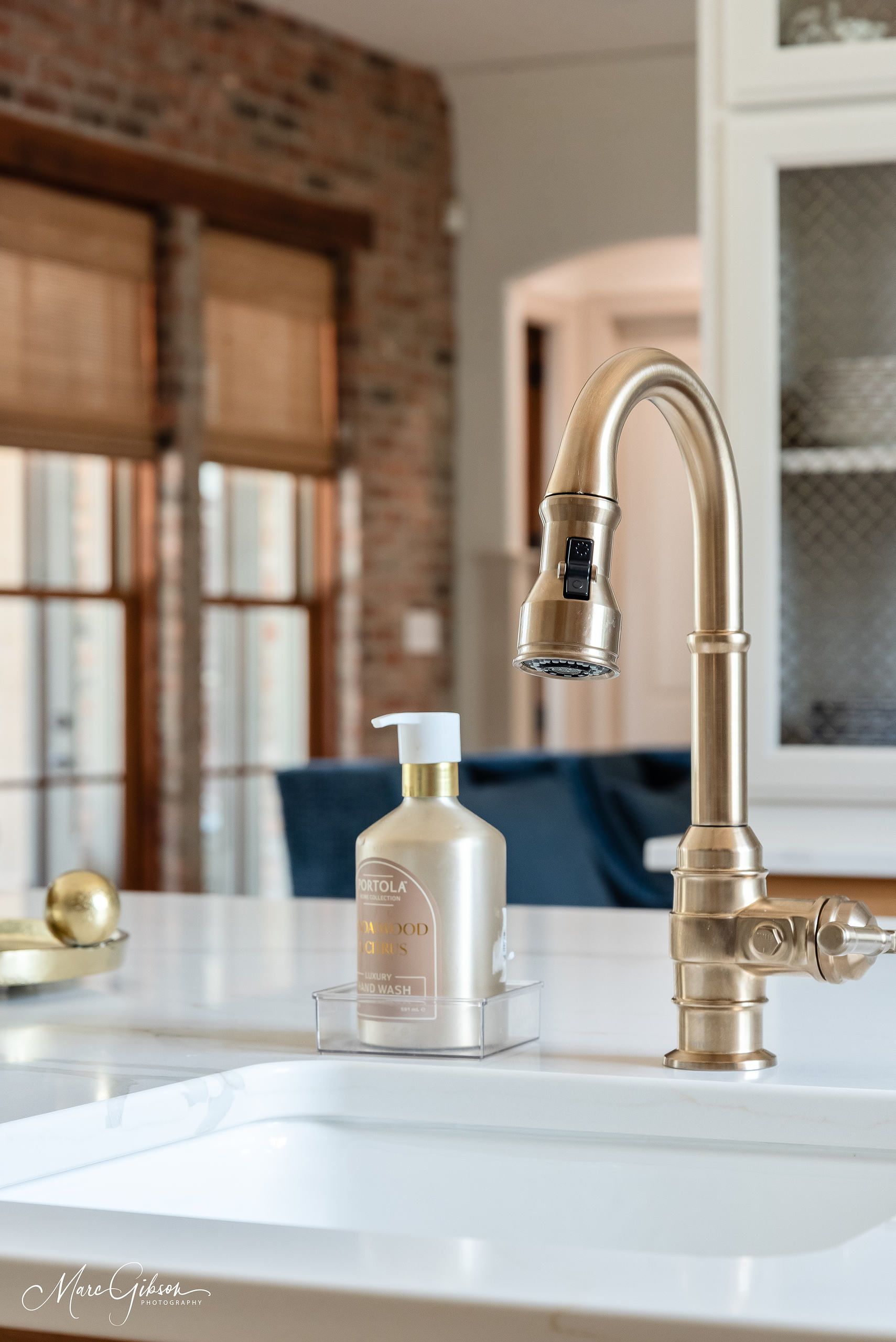 Kitchen sink with tall chrome faucet, soap dispenser, and white countertop near a brick wall and window.