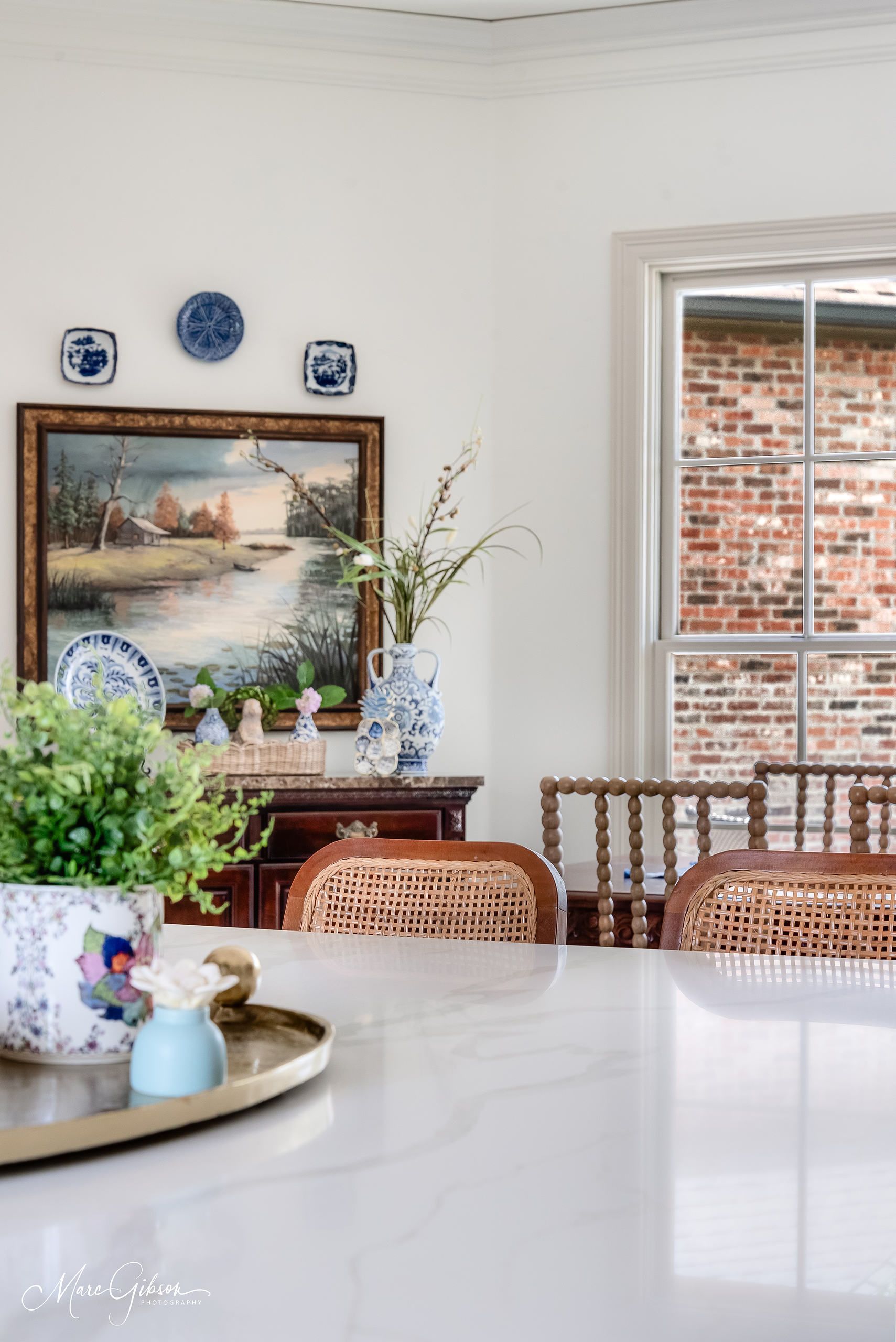 Bright dining room with a framed painting, wicker chairs, and a window beside a white marble table.