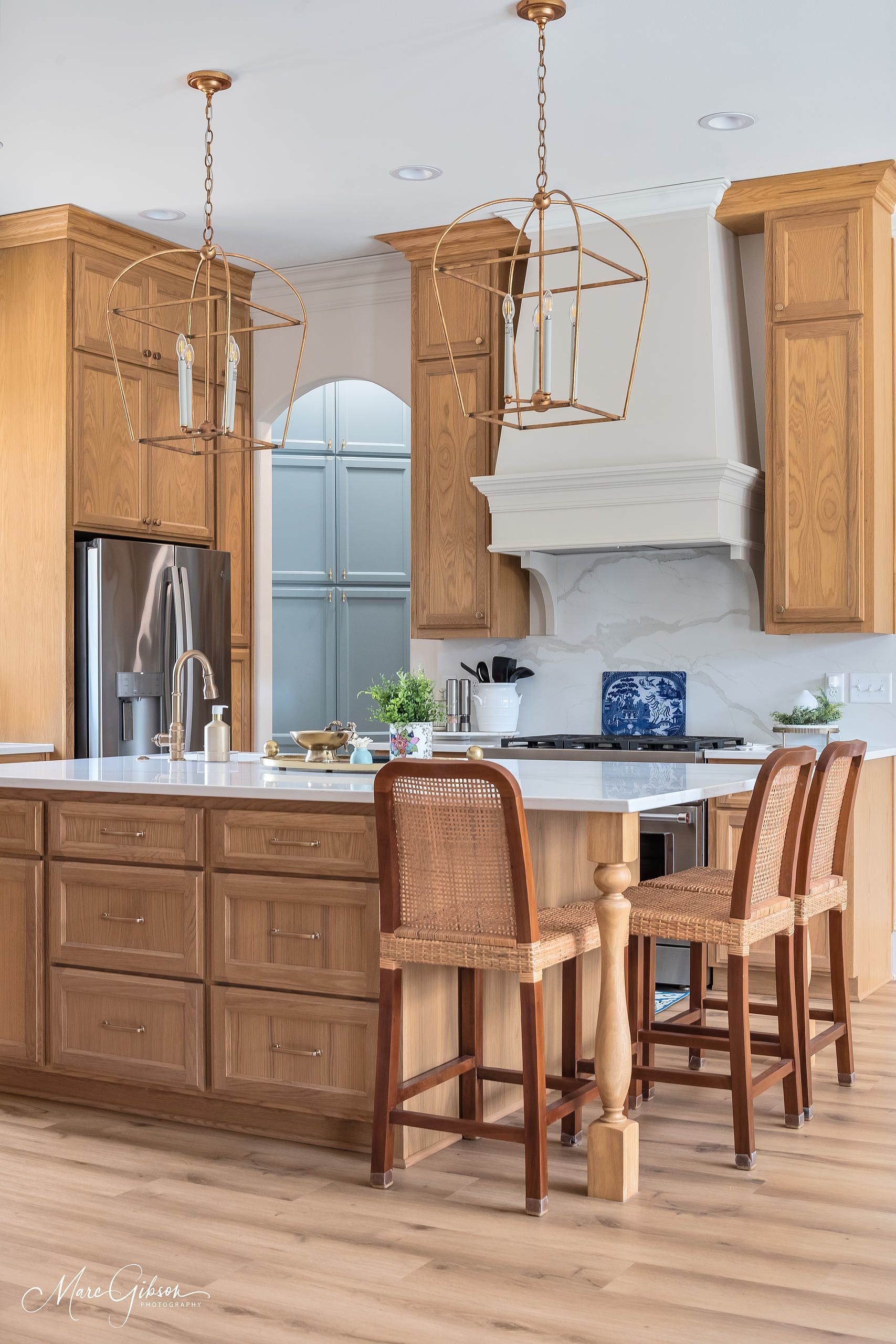Wooden kitchen with island, bar stools, stainless fridge, and white range hood under pendant lights.