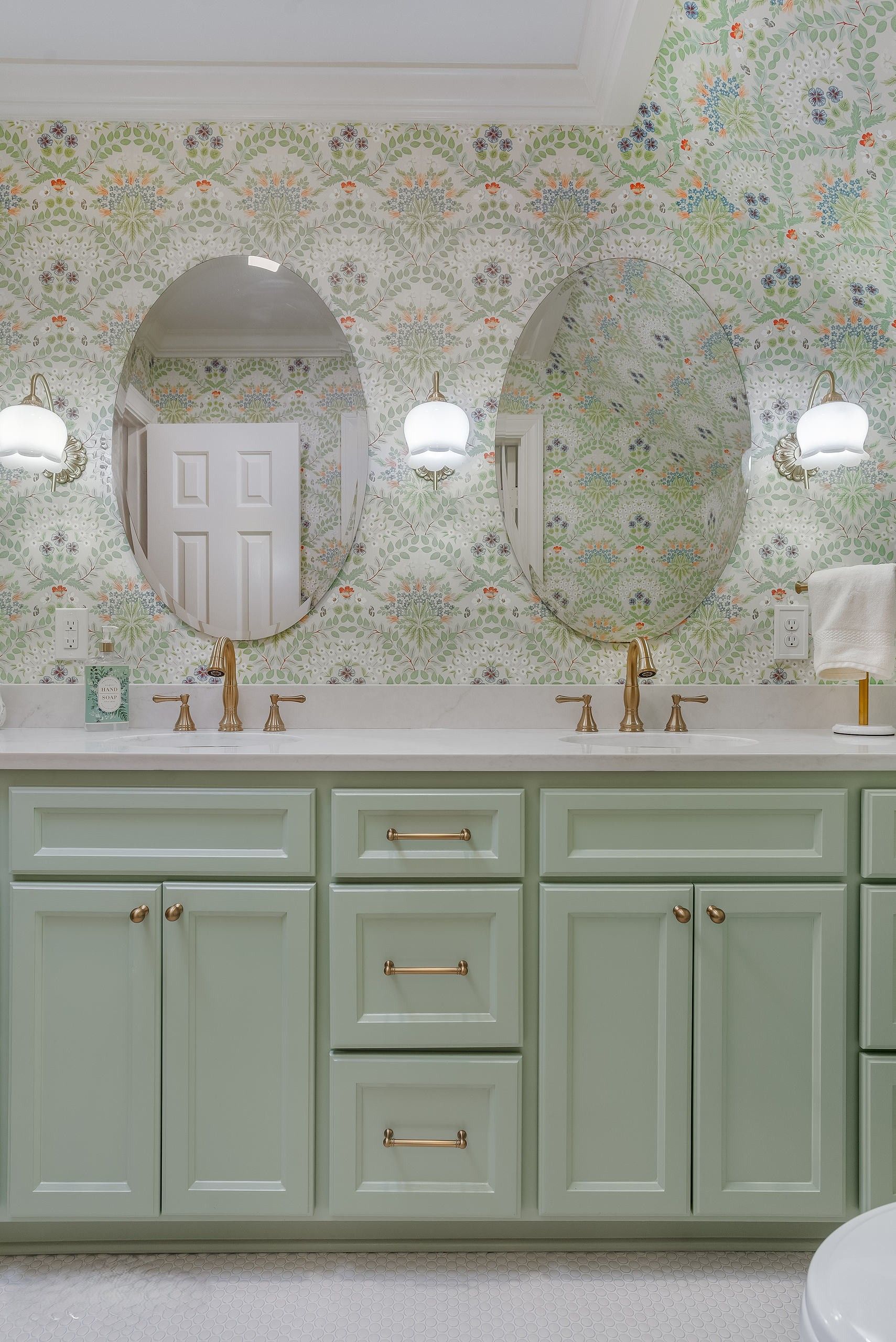 Bathroom vanity with pale green cabinets, double mirrors, and wall sconces under a patterned wallpaper.
