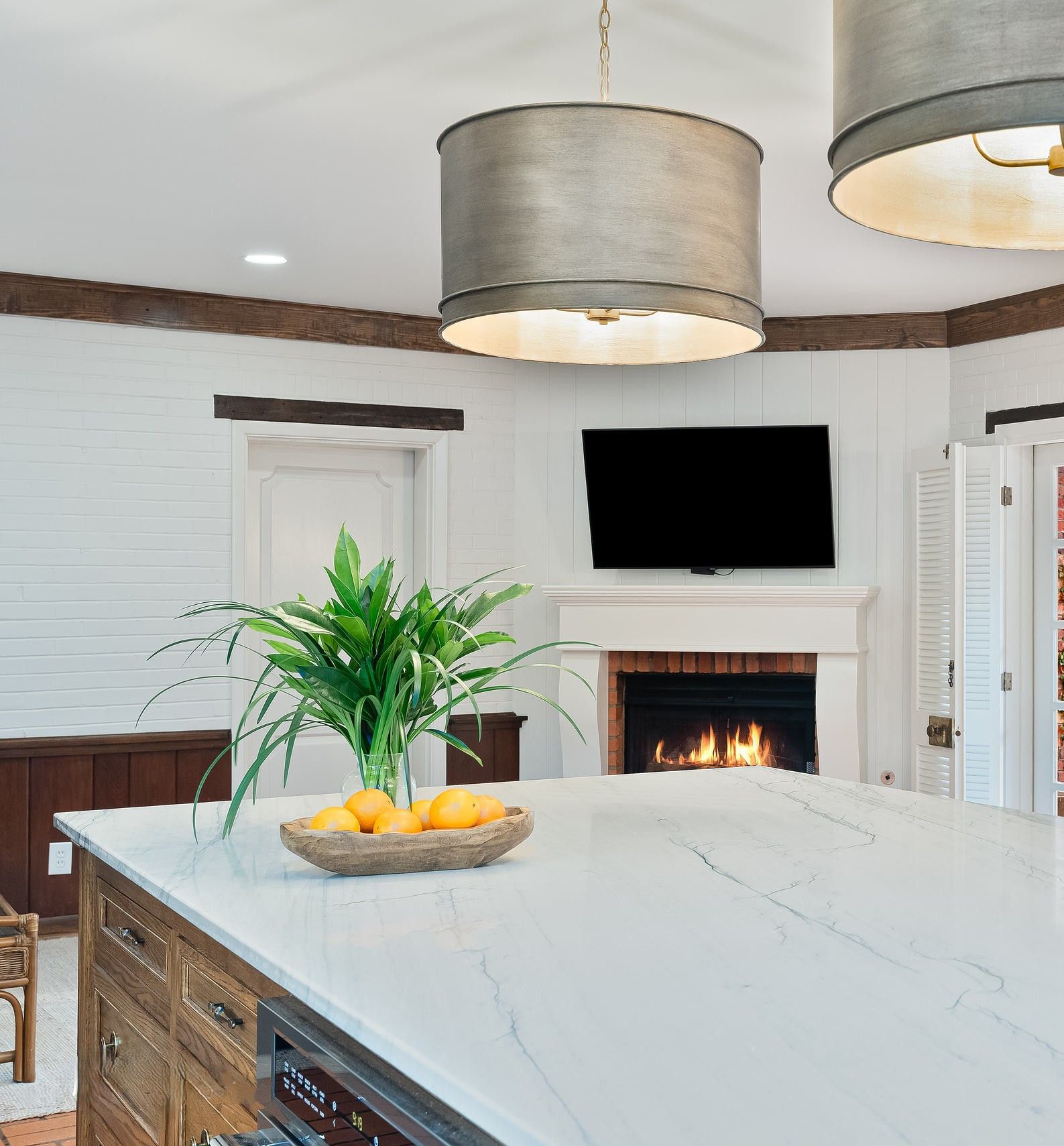 Modern kitchen with white island, fruit bowl, plant, fireplace, and wall-mounted TV