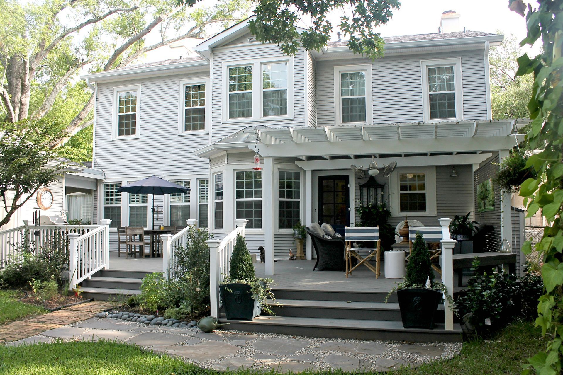 White two-story house with a wraparound porch and black rocking chairs, surrounded by trees and a lawn