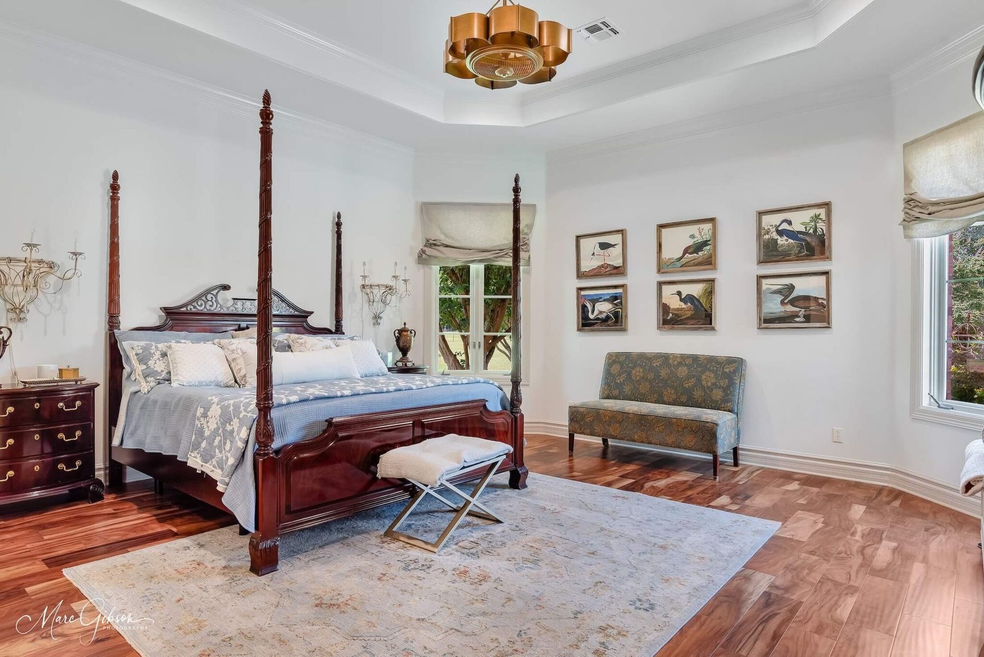 Bright bedroom with four-poster bed, dresser, rug, framed art, and large windows.