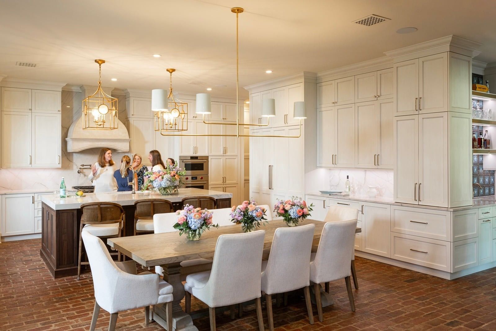 Bright white kitchen with a large dining table, pendant lights, and people gathered in the background