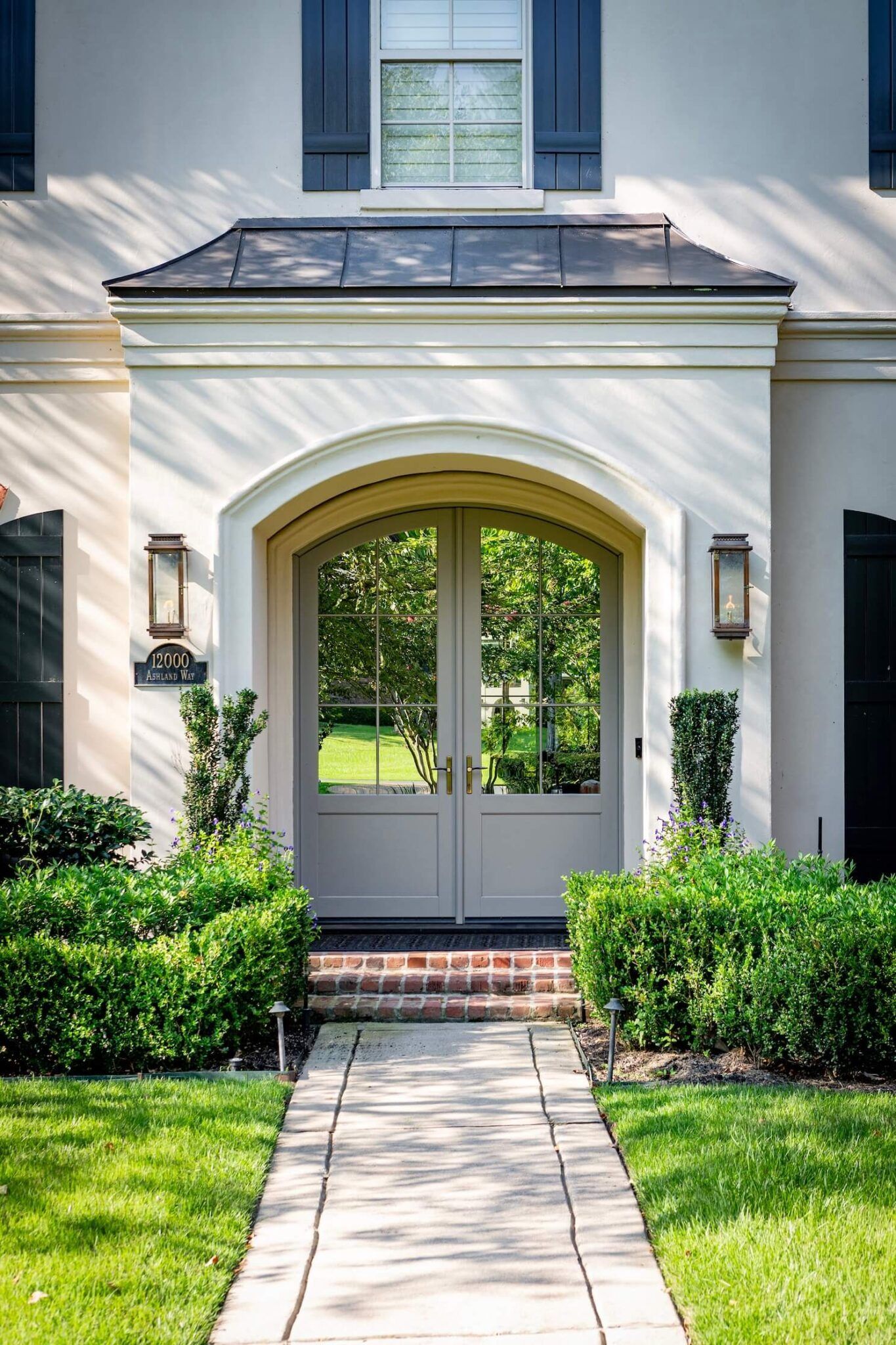 Front entrance with arched double doors, brick steps, and manicured hedges in front of a white house