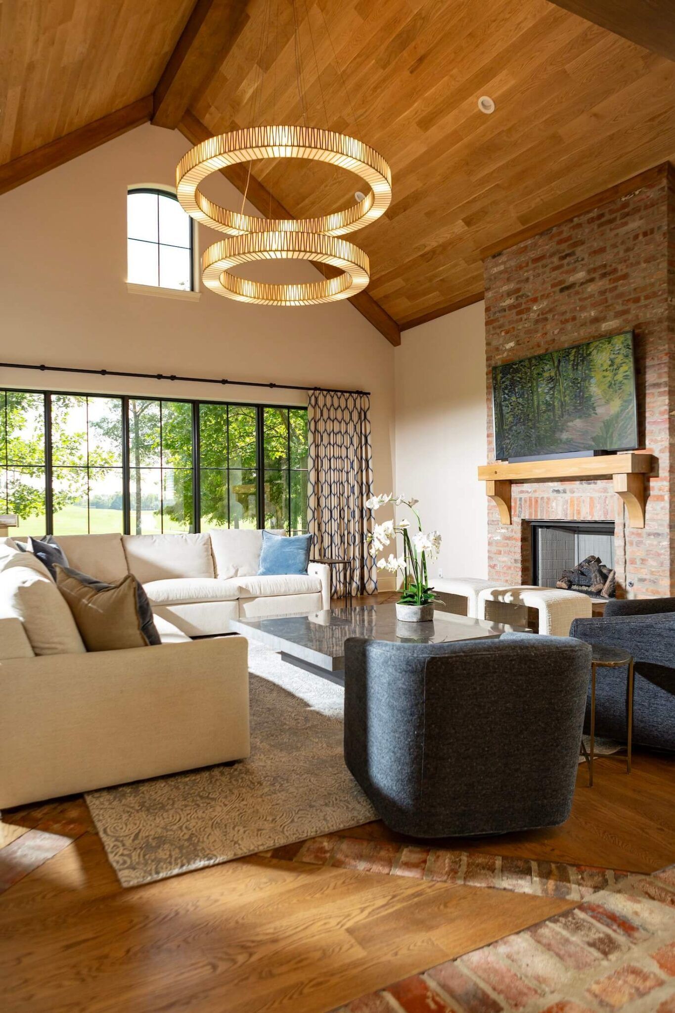 Bright living room with wood ceiling, beige sofa, black chair, large windows, and a brick fireplace