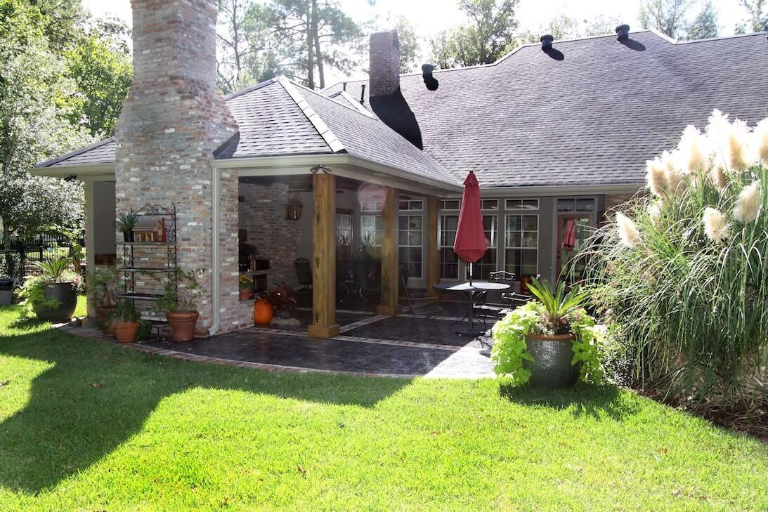 Sunlit backyard patio with stone chimney, screened porch, red umbrella, and lush green lawn