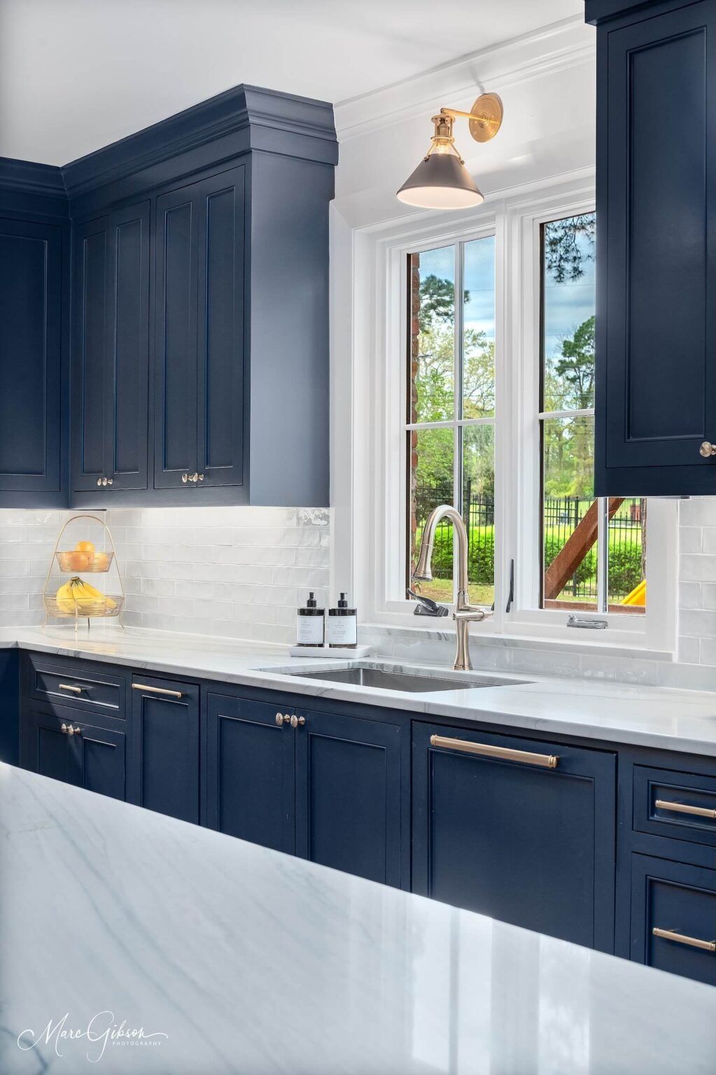 Dark blue kitchen cabinets with marble counters, white tile backsplash, and a window over the sink.