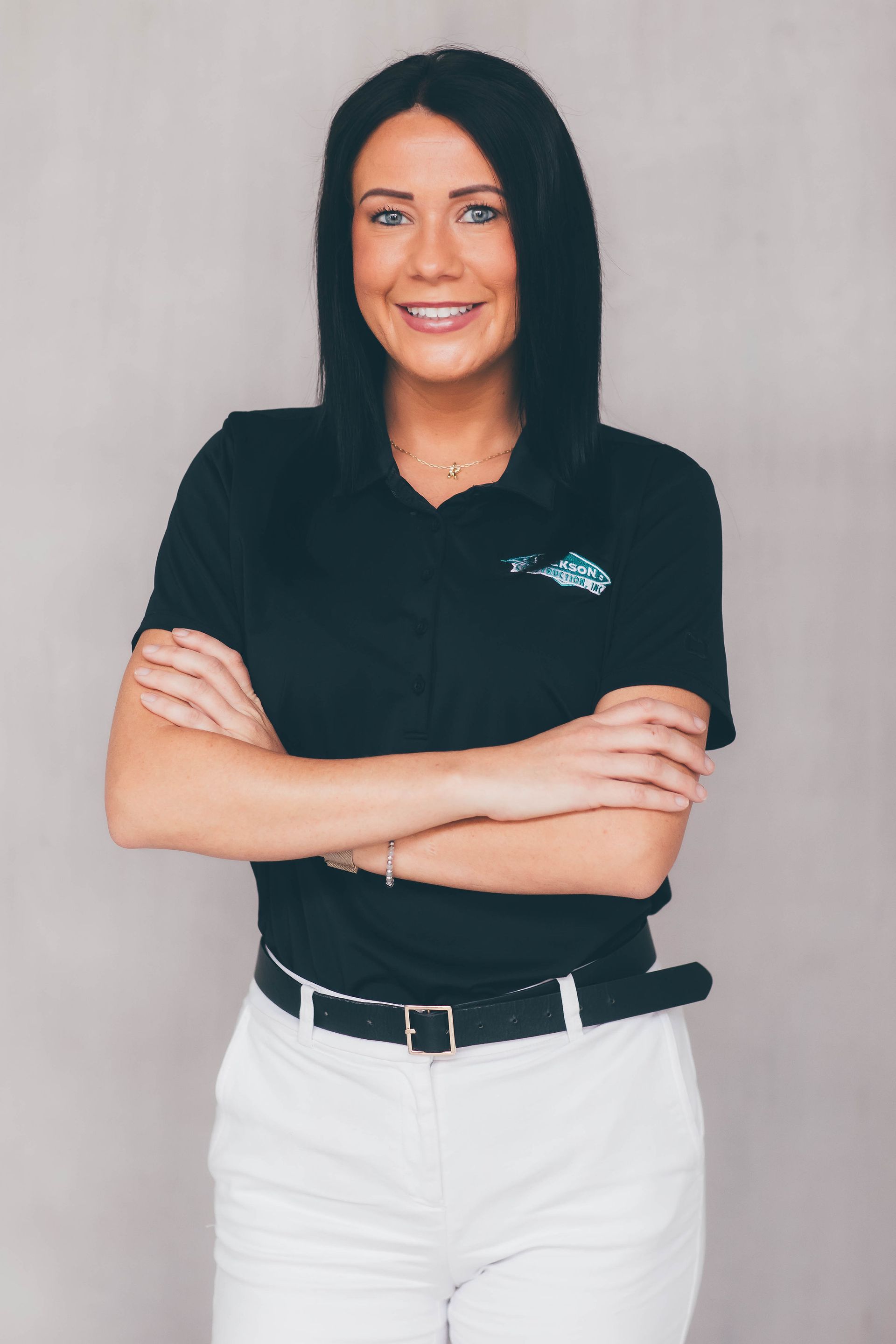 Smiling woman in black polo and white pants standing with arms crossed against a plain gray background