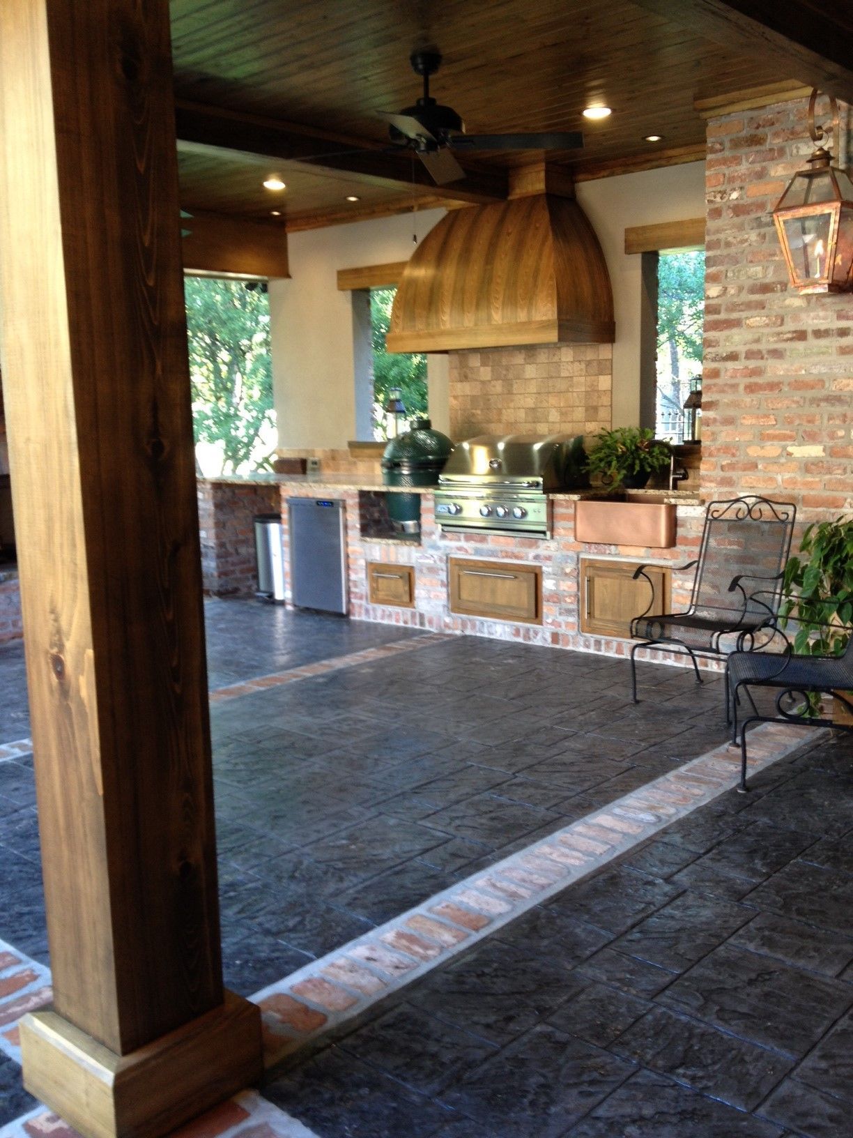 Stone outdoor kitchen with brick grill, wicker chairs, and a wooden ceiling on a tiled patio.