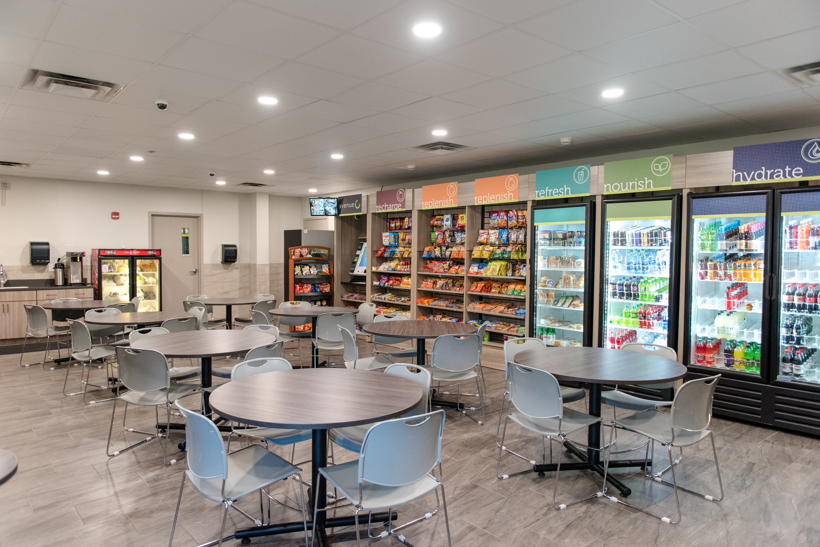 Empty cafeteria with round tables, blue chairs, vending machines, and bright ceiling lights