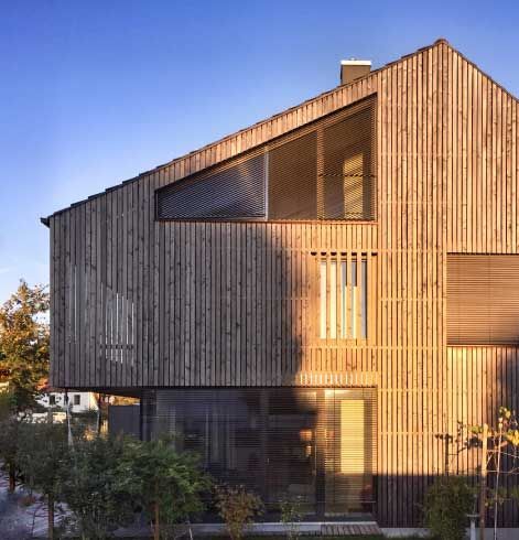 Holzhaus mit schrägem Dach und großen Fenstern; Sonnenlicht fällt auf die Holzfassade.