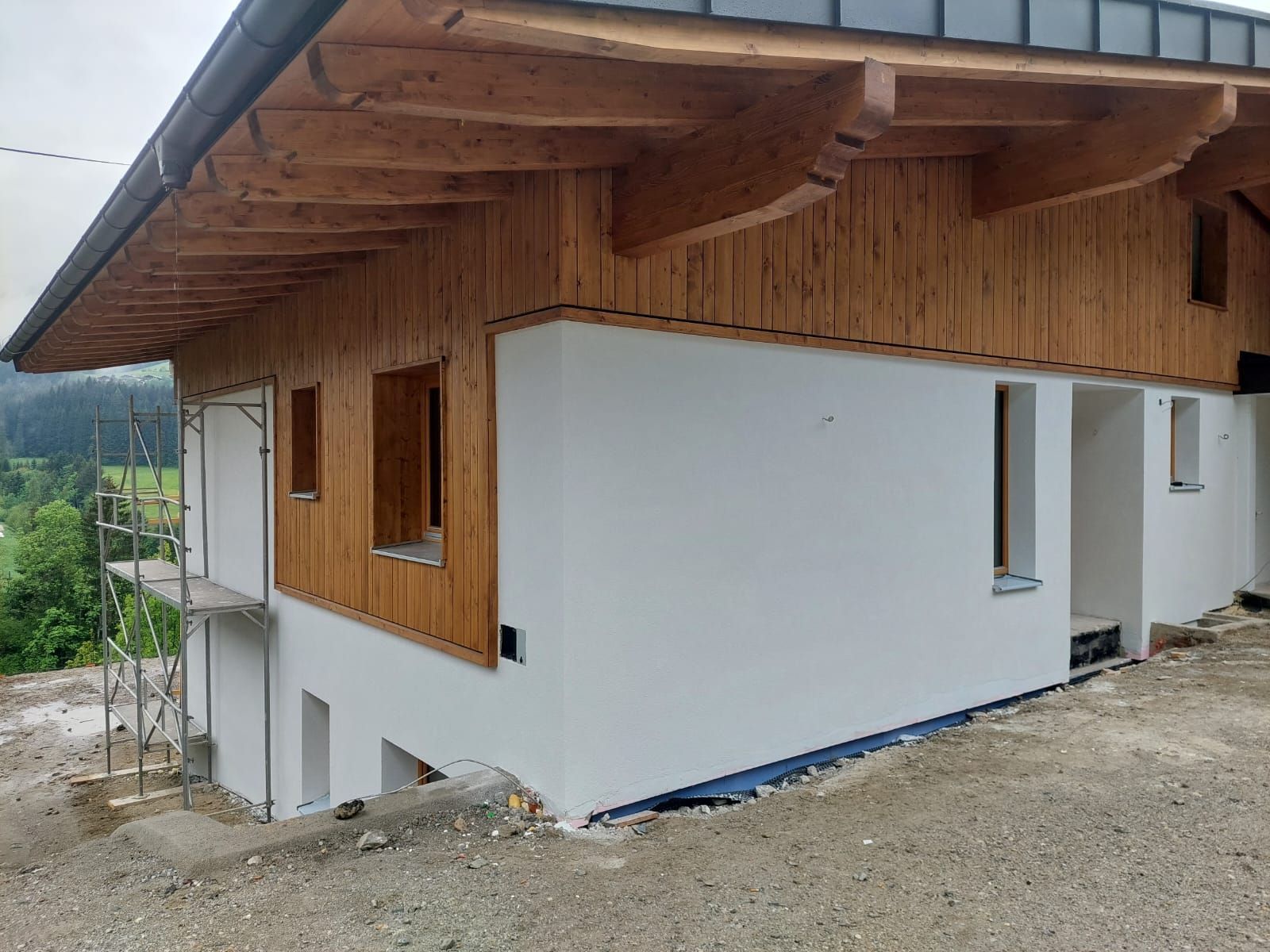 Außenansicht eines teilweise fertiggestellten Hauses mit Holzverkleidung, eingebettet in eine bergige Gegend.