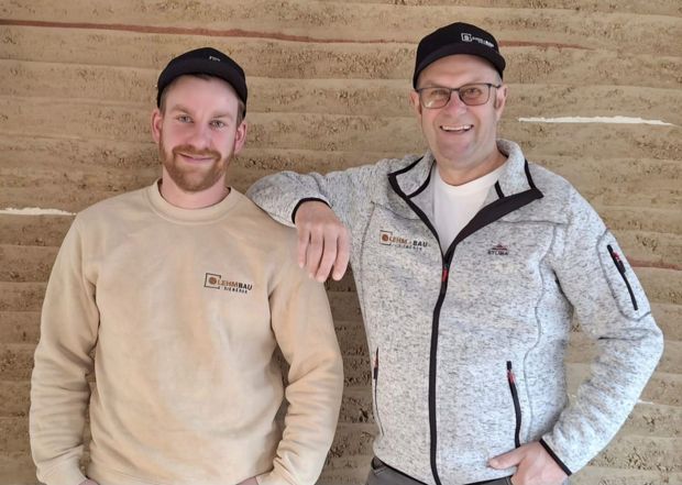 Team Lehmbau Sieberer: Zwei Männer, einer mit Bart, posieren lächelnd vor einer strukturierten Wand. 