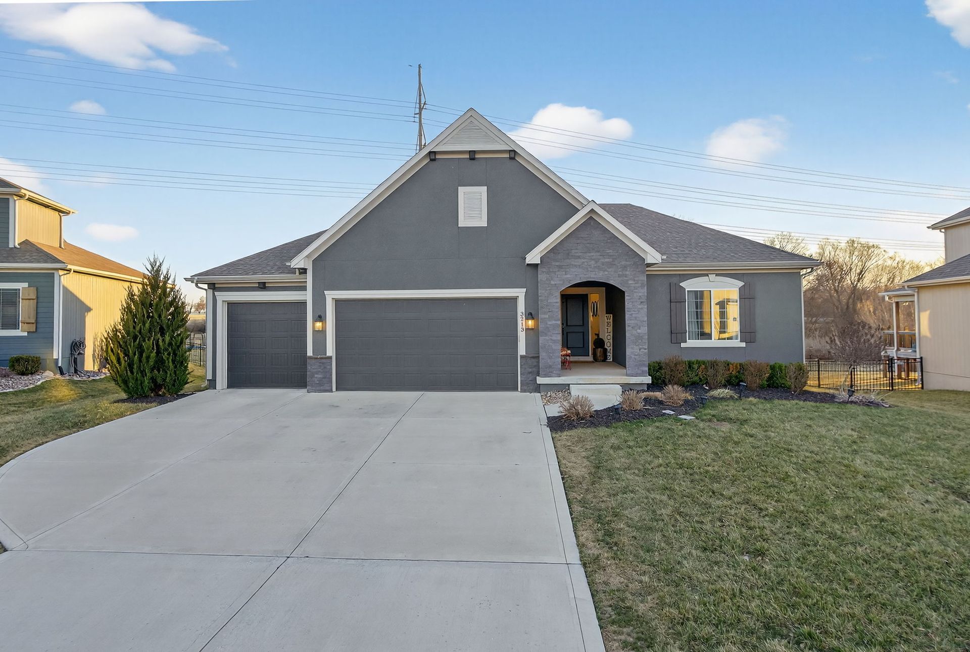 A single-story gray house with a two-car garage, concrete driveway, and a small front lawn under a blue sky.