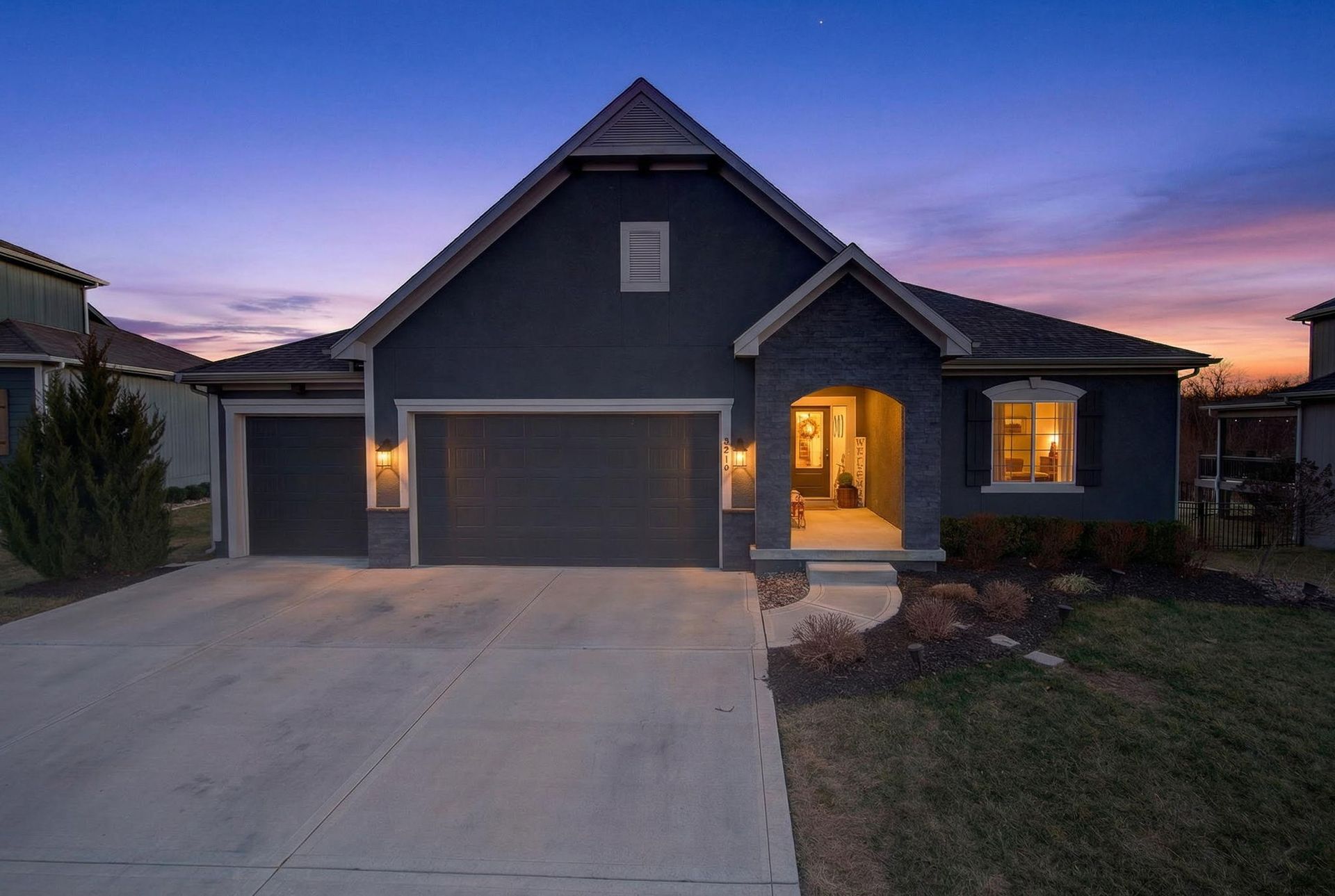 A dark blue, single-story house with a three-car garage and a front porch at dusk, under a purple and orange sunset.