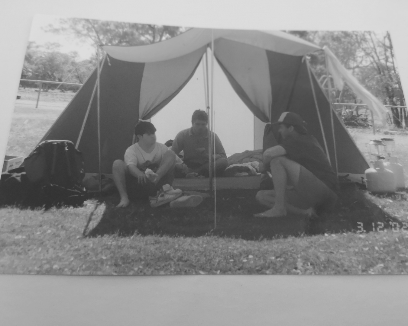 Three people sitting at the entrance of a camping tent on grass, with trees in the background.