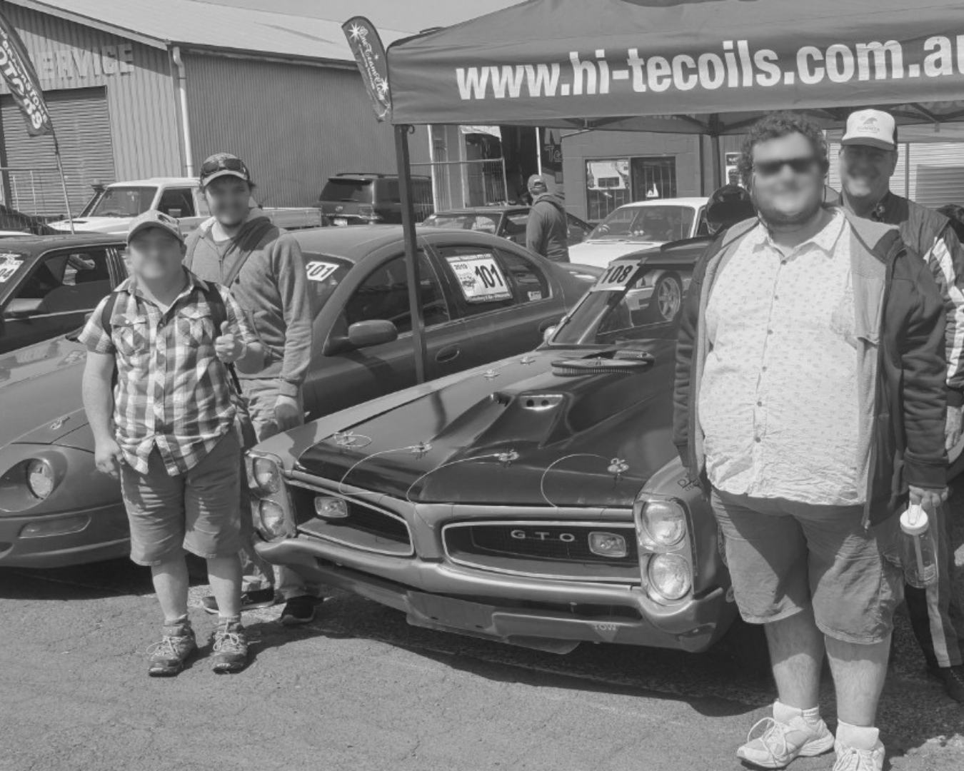 People posing beside a classic car at an outdoor auto event, with a dealership banner in the background