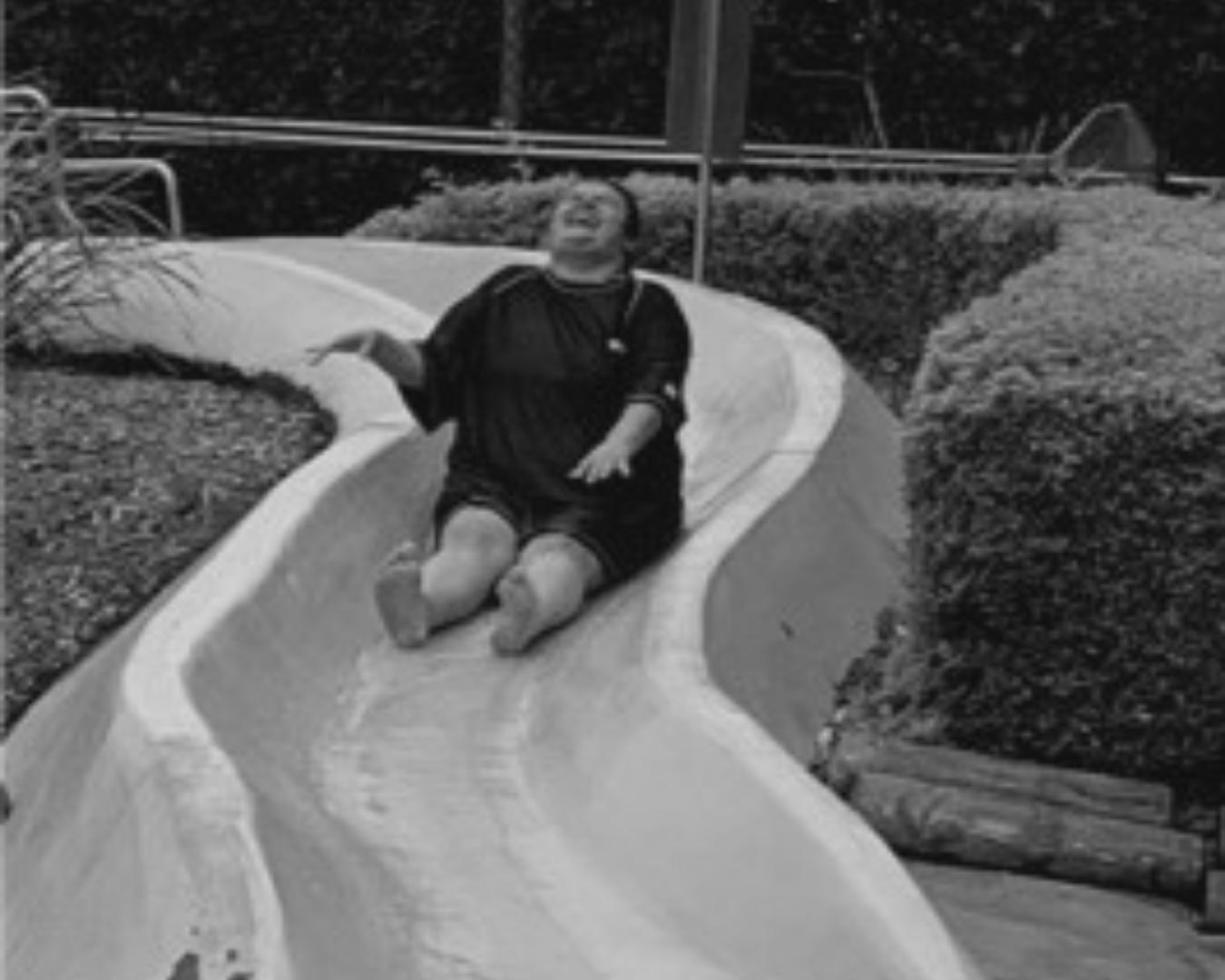 Person sitting on a curved outdoor slide, smiling and leaning back, with hedges and a fence behind them