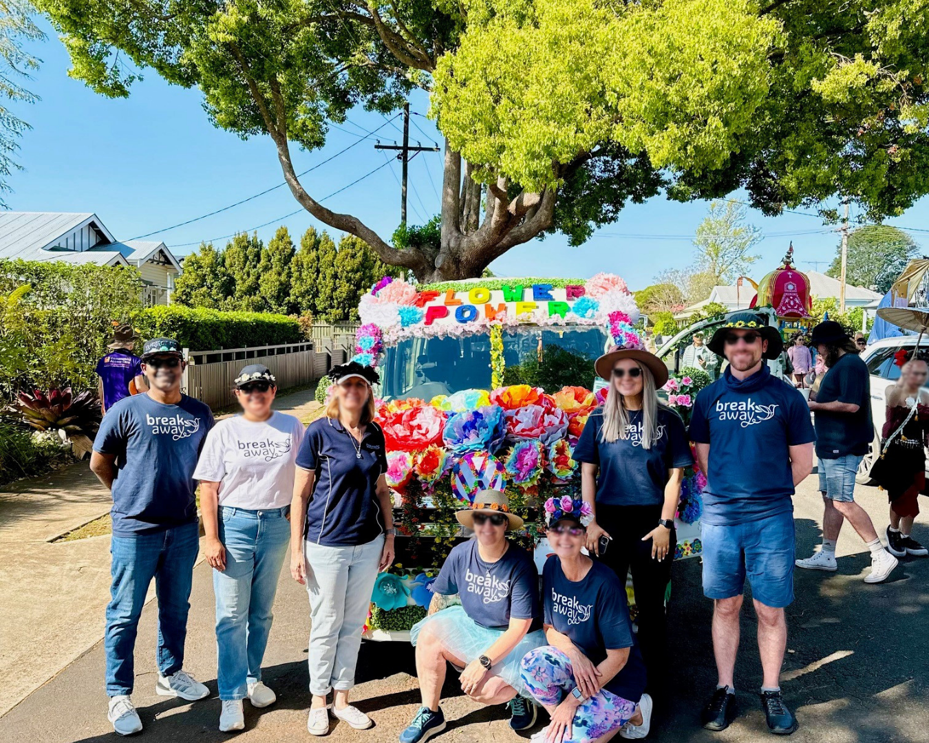 Group posing beside a colorful flower-covered parade float on a sunny street