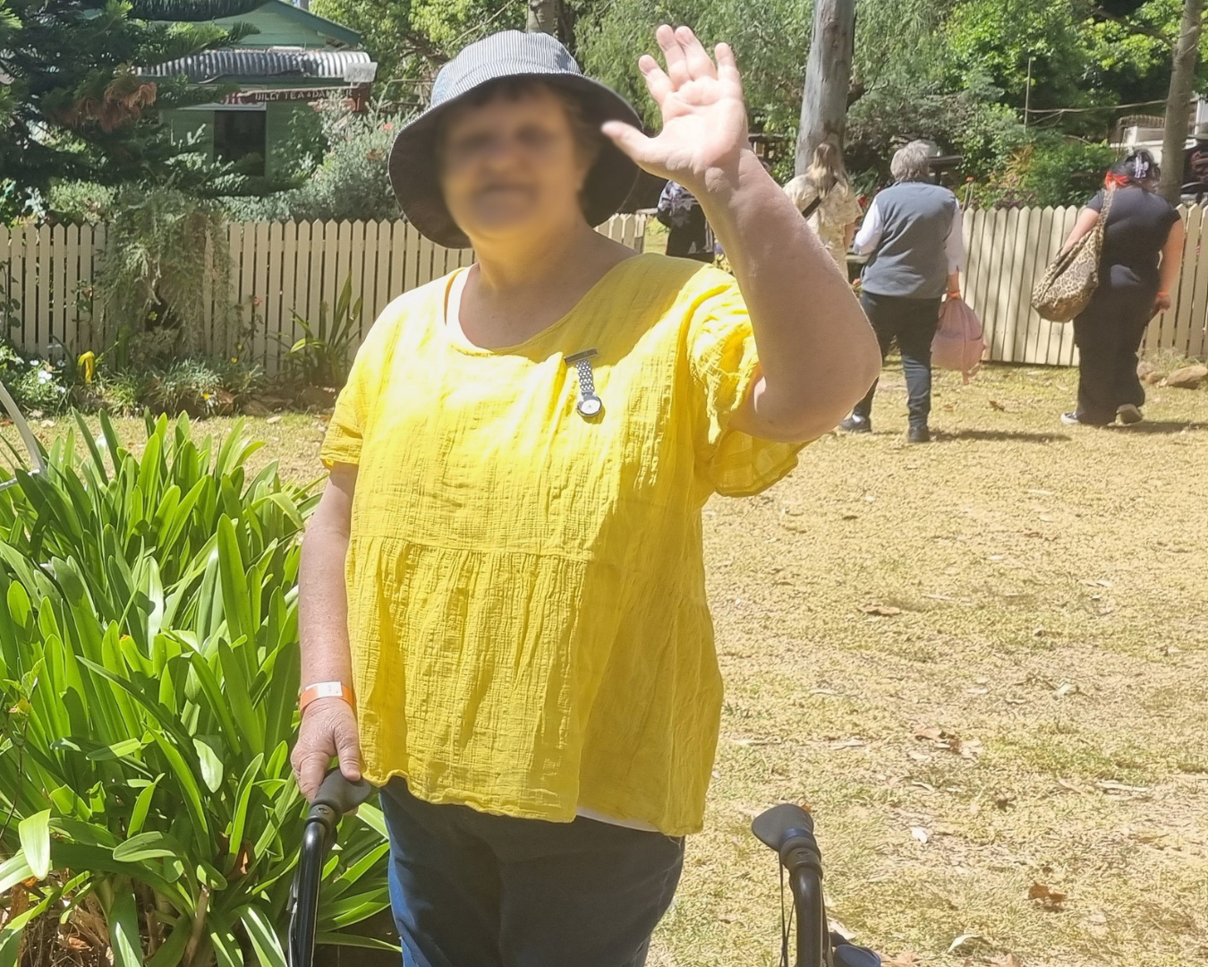 Person in a yellow top and sun hat waving in a garden on a sunny day