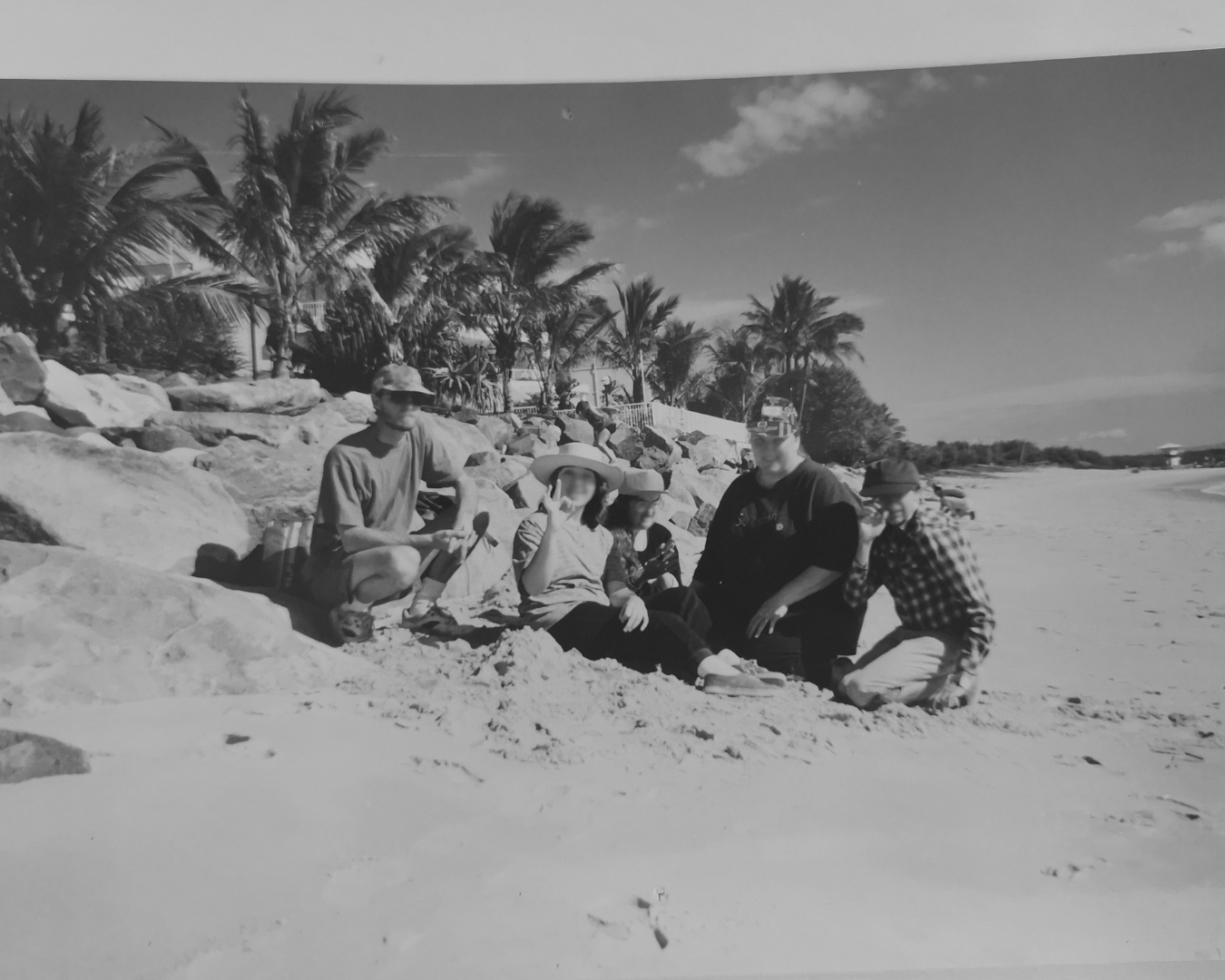 Group sitting on a sunny beach beside palm trees and rocks, with ocean in the background.