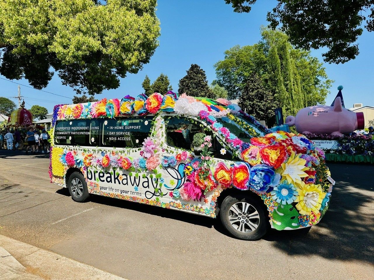 Colorfully flower-covered van parked on a sunny street, with a giant purple flower sculpture nearby.
