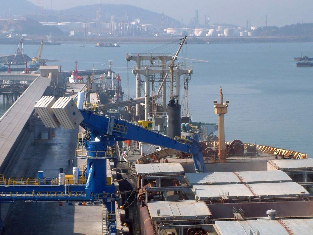 Cargo ships and cranes at a busy harbor beside a calm blue bay.