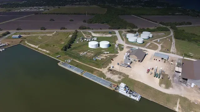 Aerial view of a riverside industrial facility with storage tanks, buildings, and a docked barge.