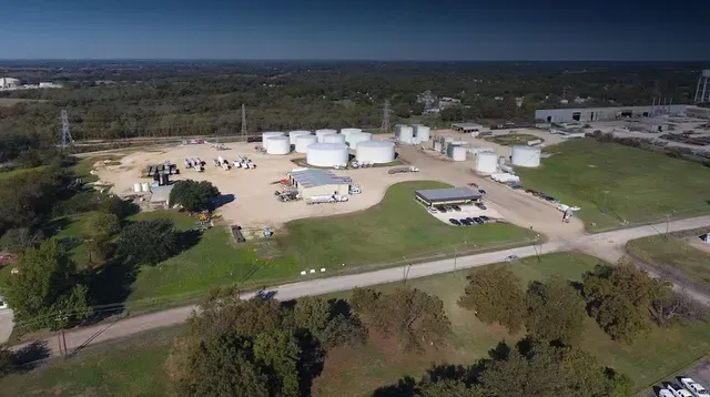 Aerial view of an industrial site with white storage tanks, buildings, and surrounding roads and fields.