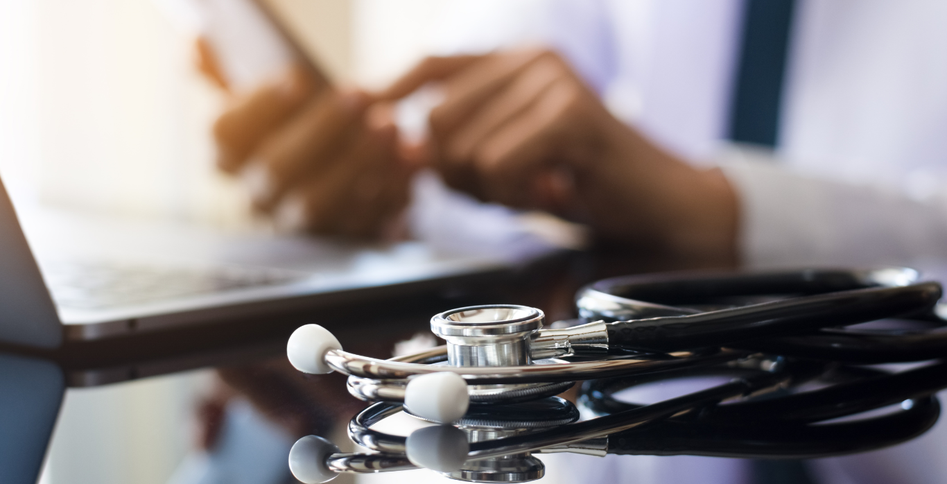Stethoscope on a desk beside a doctor writing notes on a clipboard in a medical office