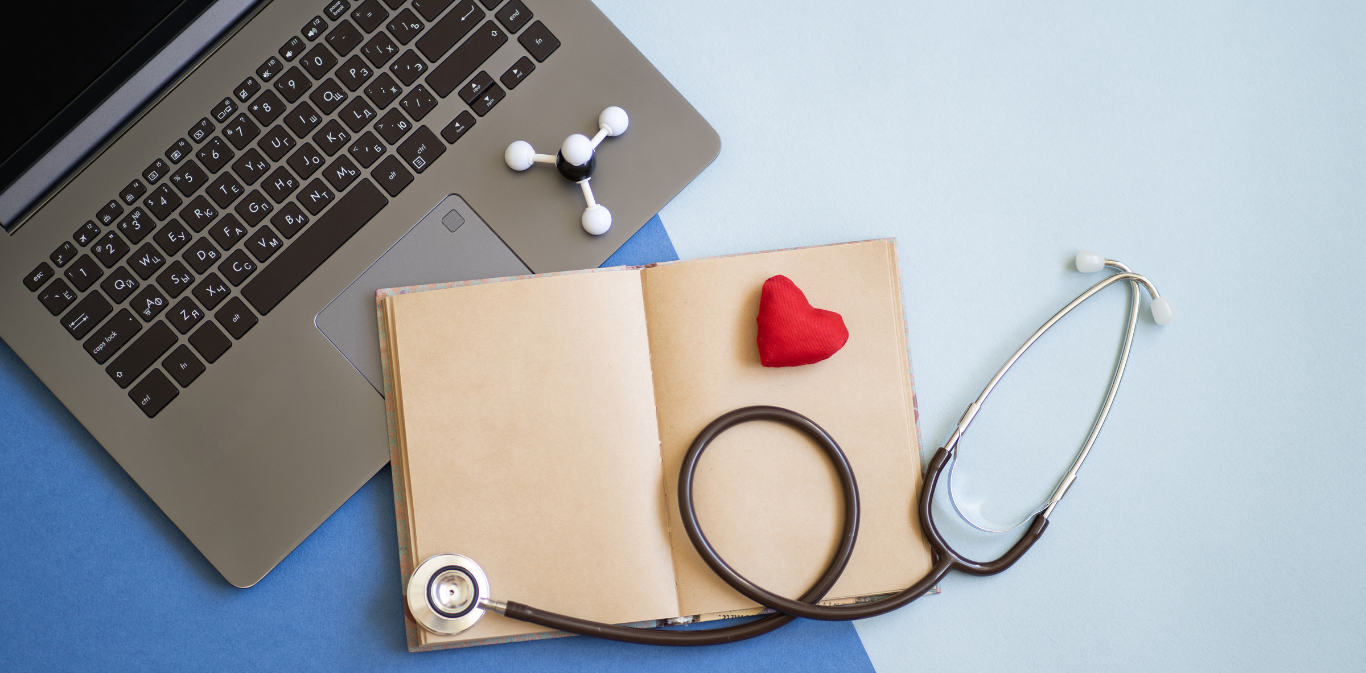 Laptop, open notebook, stethoscope, red heart, and earbuds on a desk.