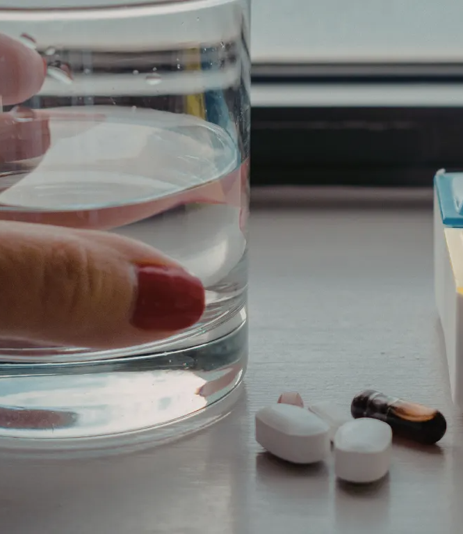 Hand resting near a water glass, with scattered white and dark pills on a table beside a pill organizer
