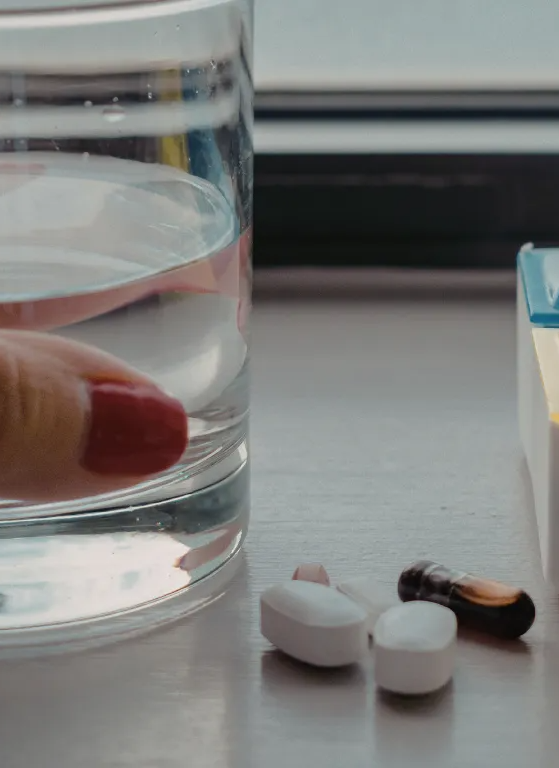 Hand holding a glass of water beside spilled white and brown capsules on a table