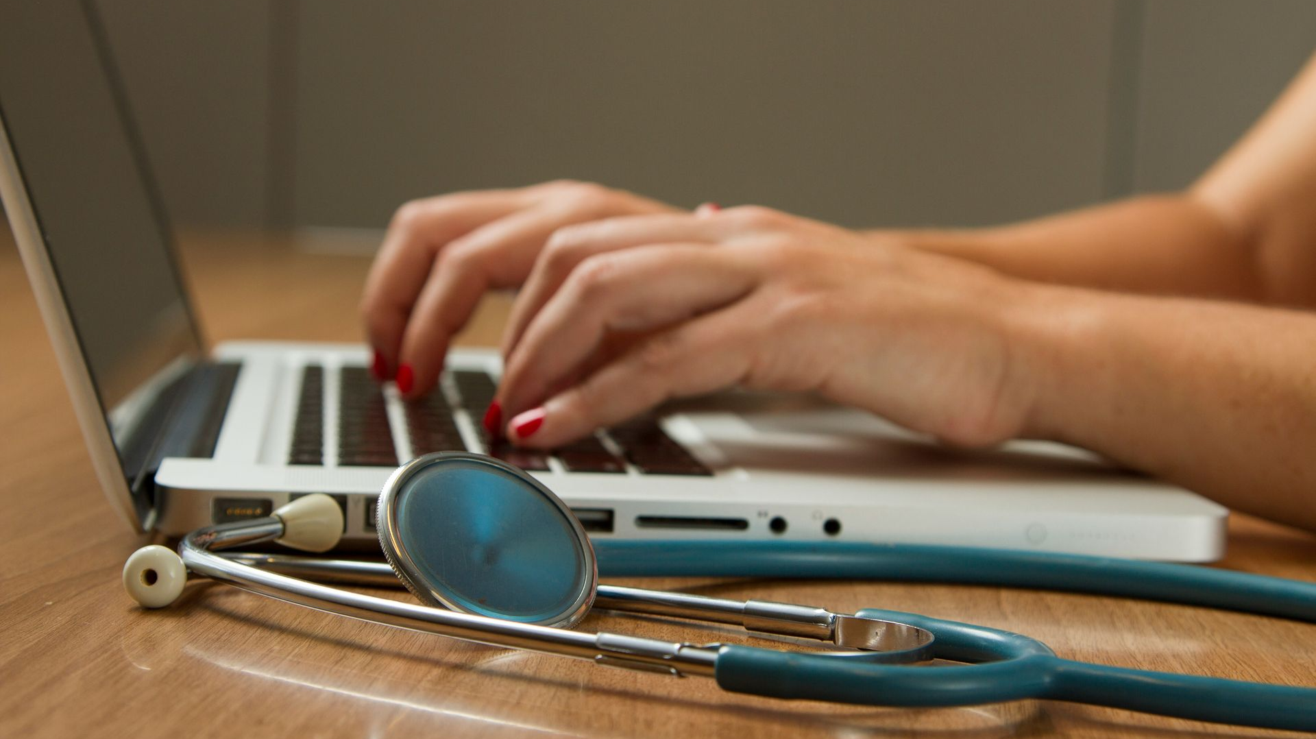 Hands typing on a laptop beside a stethoscope on a wooden desk