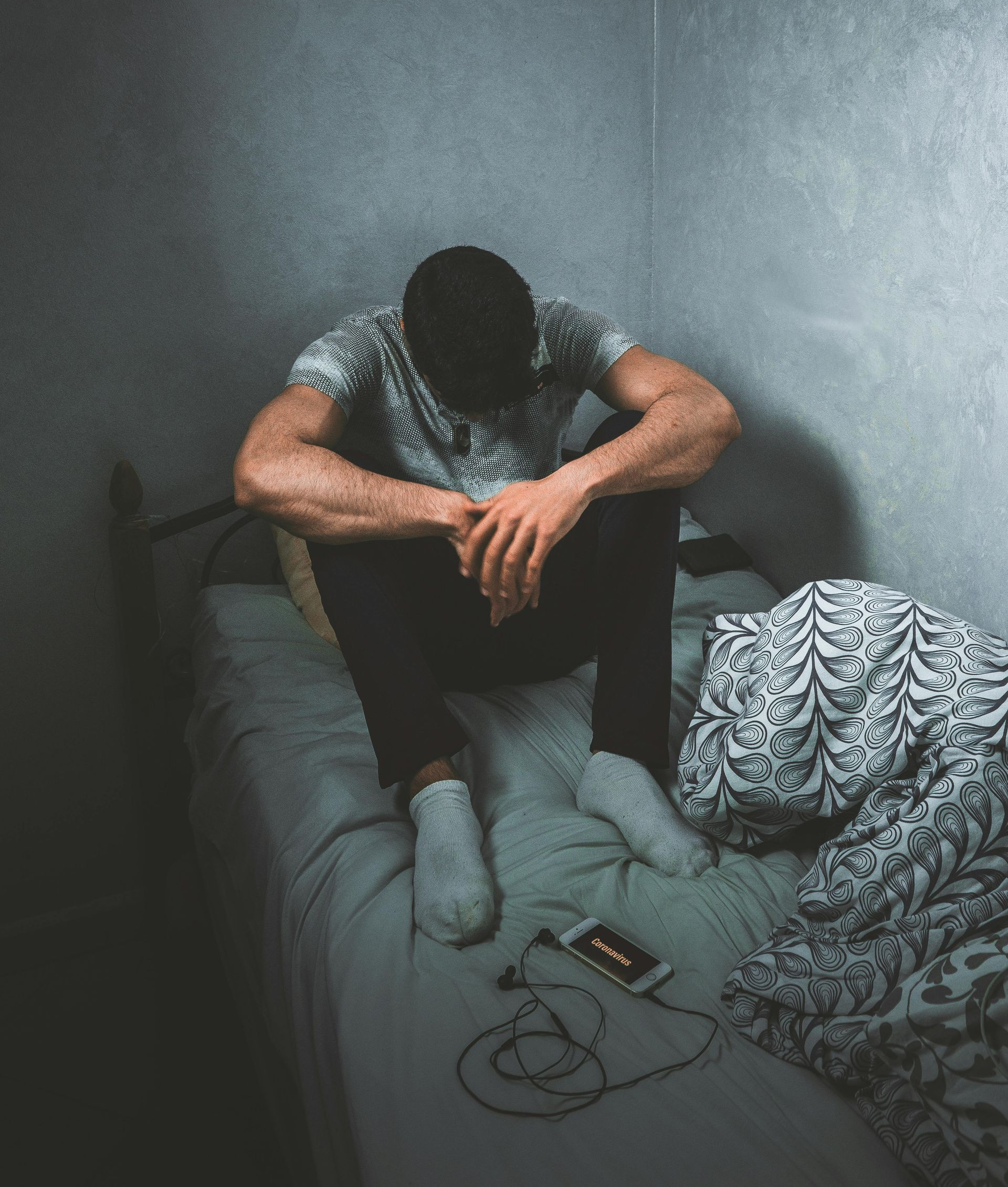 Person sits hunched on a bed in a dim room, head down beside an open laptop and tangled cords.